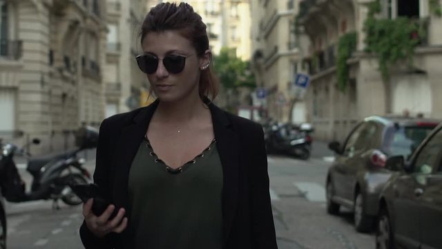 Attractive caucasian classy woman with sunglasses, freckles, piercings, black jacket and red hair having phone conversation smiling walking through the street, during sunny day in Paris. Slow motion.