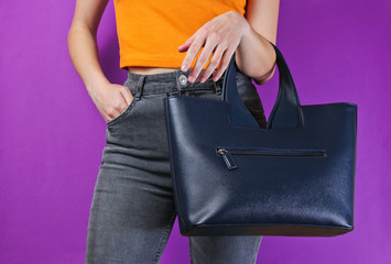 Fashionable leather bag hanging on woman's hand with orange T-shirt and gray jeans on purple background. Crop photo, studio shot