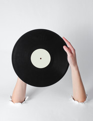 Female hands rotates a vinyl record through the torn holes of a white background. Creative art. Retro style