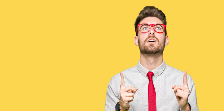 Young handsome business man wearing glasses amazed and surprised looking up and pointing with fingers and raised arms.