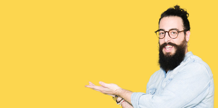Young hipster man with long hair and beard wearing glasses Pointing to the side with hand and open palm, presenting ad smiling happy and confident