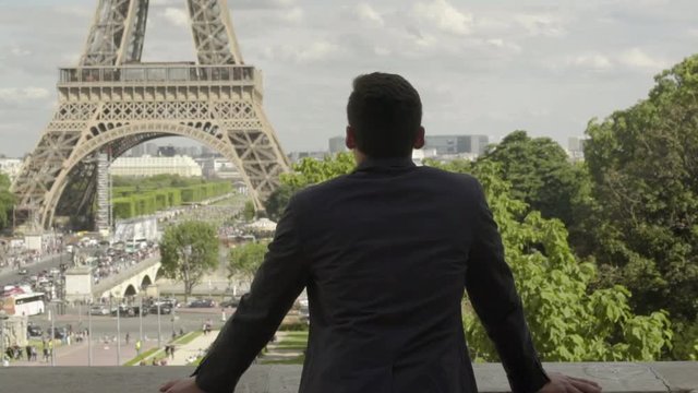 Handsome Businessman posing at the Eiffel Tower after work, with a black jacket, brown hair and tanned skin. Beautiful cloudy blue sky in Paris. 4K. Slow Motion. Peaceful, alone, view, freedom.