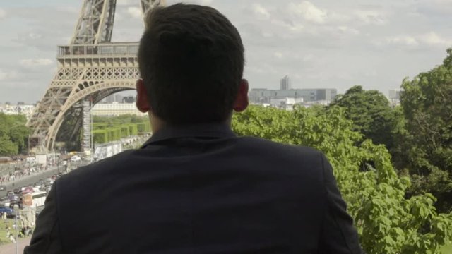 Man posing at the Eiffel Tower after work, with a black jacket, brown hair and tanned skin. Beautiful cloudy blue sky in Paris. 4K. Slow Motion. Peaceful, alone, view, freedom, success, pan to right.
