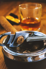 Vertical photograph of a cigar with a guillotine in the ashtray in the foreground. Against the background in defocusing a lighter and a glass with alcohol on the table