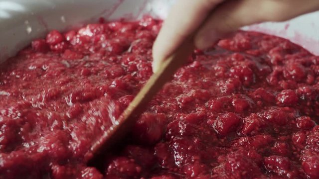 Woman stirring raspberry jam in an enameled bowl with a wooden spatula.