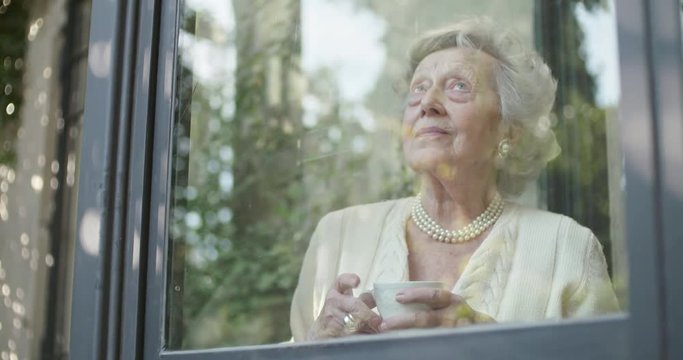 Senior grandma woman enjoying drinking cup of tea or coffee near window looking outside.Beautiful white hair elderly grandmother portrait at home.View from garden reflections. 4k slow motion video