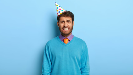 Candid shot of irritated man wears party cone hat, feels angry beacause of spoiled holiday,...