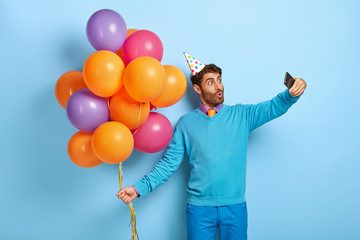 Fototapeta na wymiar Surprised handsome birthday man makes selfie portrait on cell phone, poses near colored balloons, wears blue sweater and trousers, enjoys celebration. Guy has fun on party. Congratulation concept