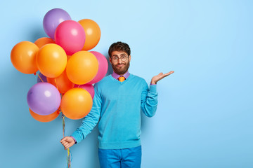 Confused hesitant male holiday maker raises palm with puzzlement, tries to make decision, holds multicolored bunch of air balloons, doesnt know whether all guests has come, wears blue clothes