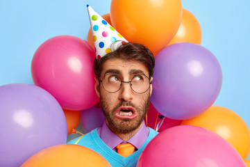 Photo of unhappy Caucasian man with miserable facial expression, looks unpleasantly aside, forgets to buy birthday cake, wears round eyewear, makes picture in photozone with colored balloons.