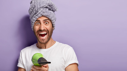 Overjoyed young man dries hair after taking shower, blows air with hair dryer, smiles broadly, wears white t shirt, towel, going to make hairstyle by himself, stands indoor against purple background