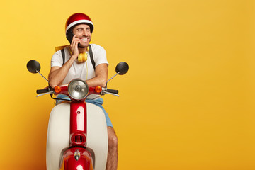 Happy man courier poses on fast motorbike, talks via cell phone during rest, carries rucksack,...