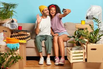 Joyful pleased girlfriend and boyfriend make selfie in their new apartment, waves in smartphone screen, have video call with friends, sit on couch with belognings. Young couple move into bought house