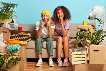 Lovely spouses of different race sit closely on comfortable sofa, being happy apartment owners, have to bring house in order after relocation, unpack boxes, rejoice successful expensive purchase