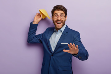 Handsome glad smiling banker throws paper plane makes wish, stretches hand, wears round spectacles, elegant formal suit, isolated on purple background. Business, achievement and success concept