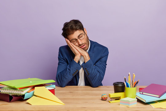 Tired overworked businessman sleeps at desktop, leans at pressed palms, works long time, busy preparing financial report, dressed in formal apparel, isolated on purple wall. Exhausted sleepy economist