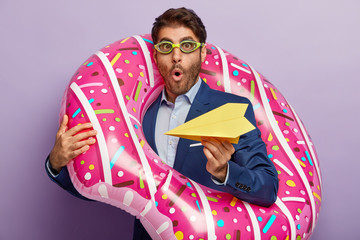 Shocked male banker holds yellow paper plane, dreams in office about resting on seaside, carries...