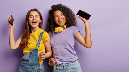 Two happy women dance with joy, listen music on smartphones, have fun together, dressed casually, move against purple background, free space on right side. Mixed race friends have perfect mood