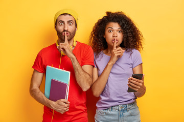 Image of multiethnic teenage friends show silence gesture, ask keep information confidential, use cell phone for chatting online, gossip about groupmates, isolated on yellow background. Omg, be silent