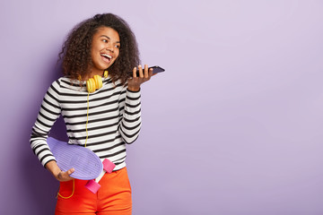 Waist up shot of smiling carefree dark skinned woman makes sound call, holds cell phone near mouth, wears headphones on neck, holds longboard, enjoys spare time, stands indoor. Skateboarding concept