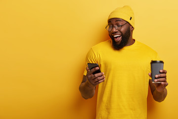 Cheerful dark skinned man uses smartphone application, watches video in internet, chats online, drinks coffee from disposable cup, wears yellow clothes, stands indoor, ignores live communication