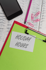 Handwriting text writing Holiday Hours. Conceptual photo employee receives twice their normal pay for all hours Clipboard sheet pencil smartphone squared note clips wooden background
