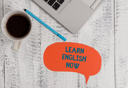 Text sign showing Learn English Now. Business photo showcasing gain or acquire knowledge and skill of english language Metallic open laptop speech bubble coffee cup ballpoint pen vintage table