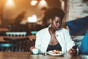 Fototapeta na wymiar An elegant young black businesswoman in a white cloak and spectacles with curly Afro hair is having lunch in a cafe with coffee and salad, holding her smartphone and scrolling feed of social networks