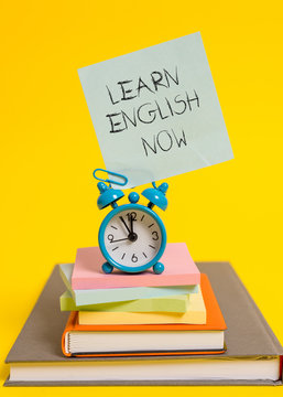 Handwriting text writing Learn English Now. Conceptual photo gain or acquire knowledge and skill of english language Alarm clock note stacked notepads old books colored background