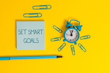 Word writing text Set Smart Goals. Business photo showcasing giving criteria to guide in the setting of objectives Metal alarm clock wakeup clips ballpoint notepad colored background