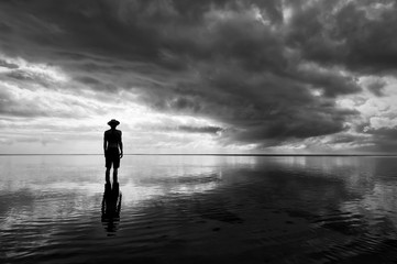 Moody monochrome silhouette of man in hat standing in calm shallow waters reflecting dramatic...