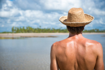 Caucasian tourist wearing a straw sun hat looking out at a bright palm-lined island beach scene,...