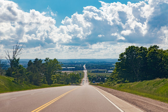 Beautiful Landscape Midday View Of Canadian Ontario Country Side Road With Cars Traffic During Sunny Day With White Clouds In Blue Sky. Busy Outdoor Country Village Street At Summer.