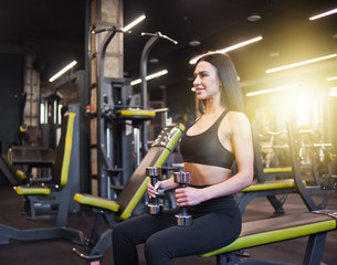 Fototapeta na wymiar Smilling young woman holds dumbbells in hands while sitting on a bench in gym