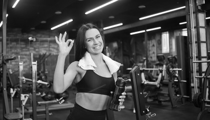 Fototapeta na wymiar Portrait of a cheerful smilling young sportswomanin in sportswear and towel around his neck, bottle of water in hand shows fingers ok sign in gym. Positive emotions, training process