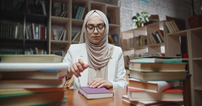 Female muslim student writes down notes in her notebook, sitting at desk covered with books and looking at camera - modern islam, student lifestyle concept close up 4k portrait