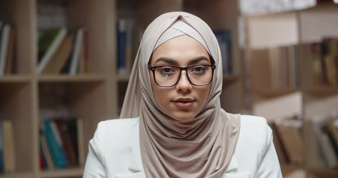 Portrait of female muslim student, wearing hijab, who is at campus library and confidently looking at camera - modern islam concept 4k