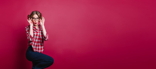 Inspired barefooted girl in glasses standing on one leg in studio. Indoor photo of enthusiastic young woman in checkered shirt posing on claret background.