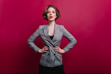 Winsome business lady with short haircut posing in studio. Photo of confident girl in tweed jacket isolated on claret background.