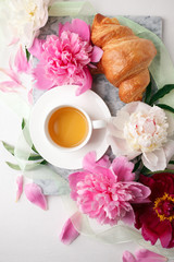 cup of tea herbal, croissant and peony buds on marble plate on white table. Beauty breakfast composition, celebration seasonal holiday feminine background