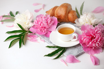 cup of tea herbal, croissant and peony buds on marble plate on white table. Beauty breakfast composition, celebration seasonal holiday feminine background