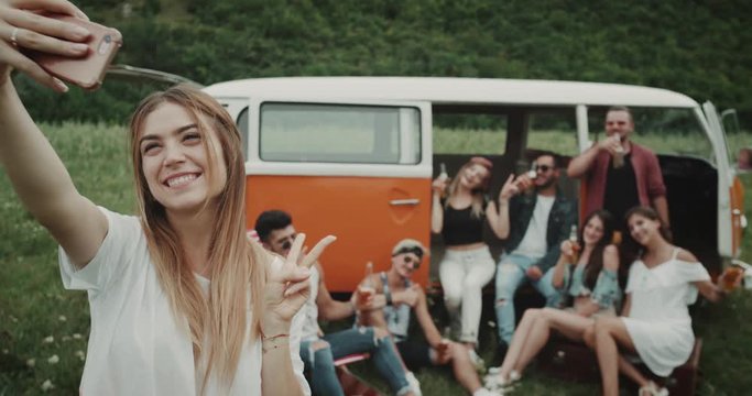 Lady taking selfie from phone with her big group of friends at the picnic in the middle of field, behind of a retro van