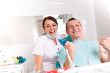 Fototapeta premium Dentist finishing successful teeth restoration, black haired doctor, in green mask, giving to happy patient mirror, beautiful long-haired woman looking at pristine white teeth with great pleasure