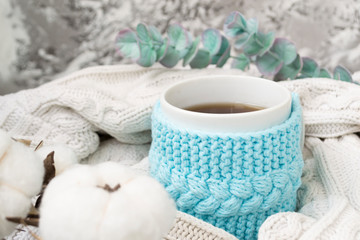 Fototapeta na wymiar White mug with tea in a knitted blue frame and wrapped in a white knitted plaid. In the background, sprigs of eucalyptus and cotton. Breakfast in bed. Cozy.