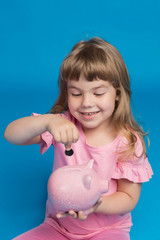happy girl, in pink clothes, with a pink piggy bank, smiles and puts a coin in the piggy bank, finance and accumulation concept