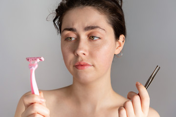 A young pretty woman holding a pair of tweezers and a razor, not knowing how to remove excess hair on her face.The concept of getting rid of unwanted hair