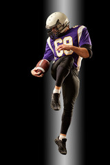 Fototapeta na wymiar American football player holding a ball while jumping away from a strike. Black background, copy space. Violet with white sportswear.