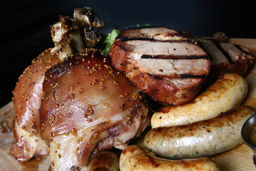 Assorted grilled meat. Knuckle, steaks, homemade sausages on a wooden board. Close up on a black background.