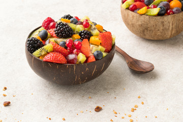 Colourful fruit salad in a coconut bowl and wooden spoon white  stone surface