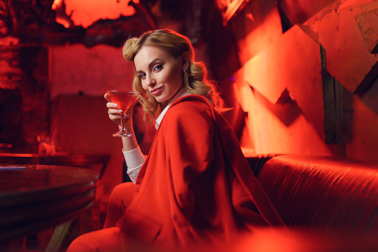 Image of happy blonde woman in red jacket looking at camera with cocktail in her hand in nightclub
